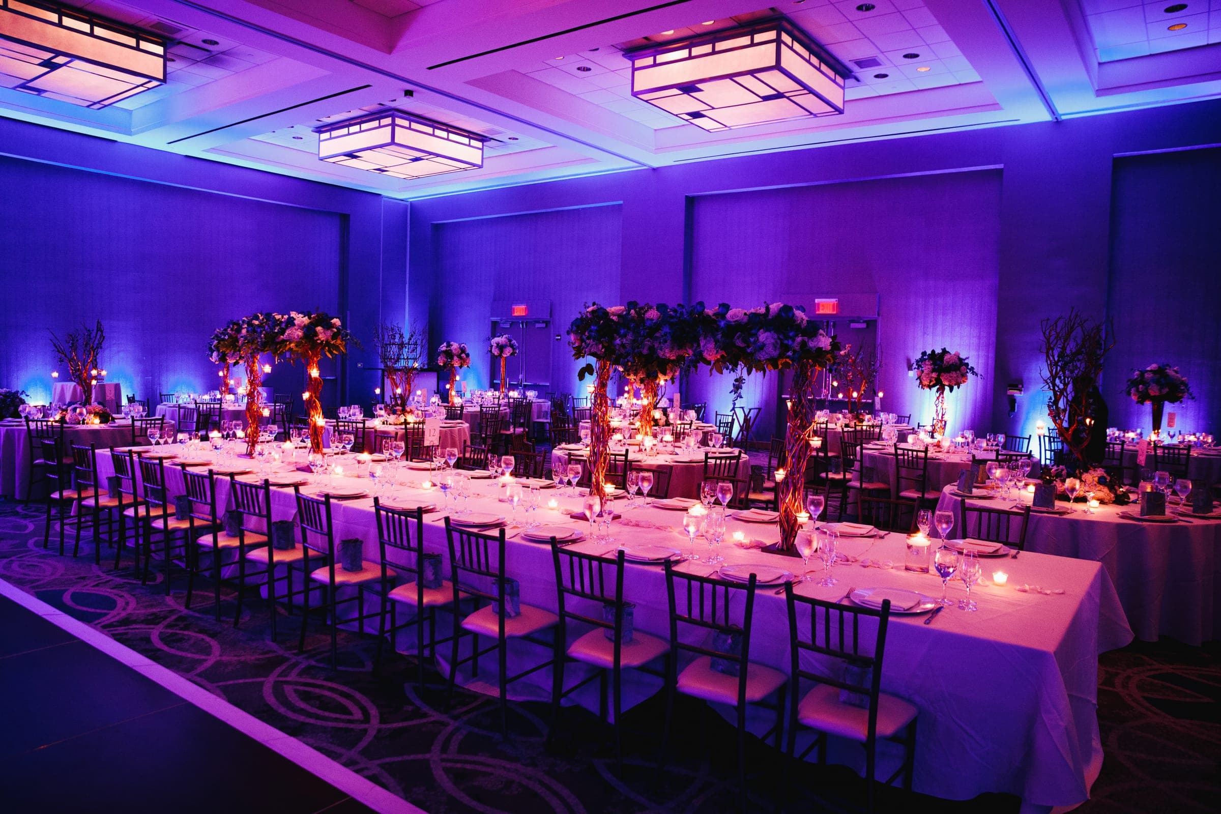 A formally set banquet hall with long tables, floral centrepieces and purple uplighting