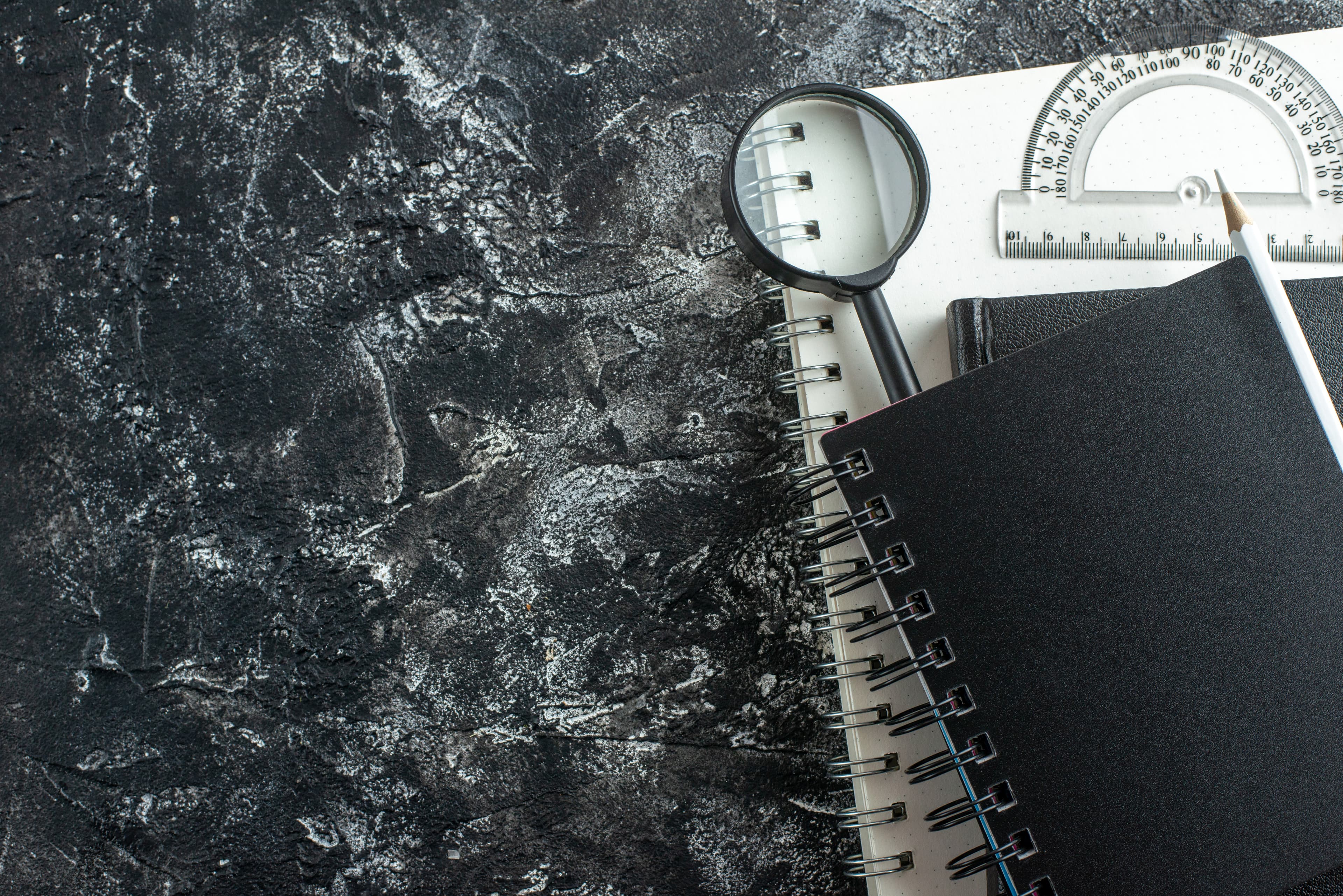 A notebook, magnifying glass, ruler and pencil laid out on a dark textured desk